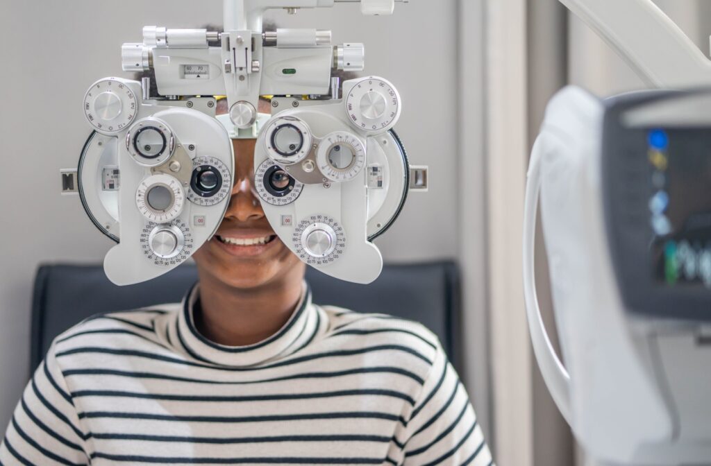 Teenager in striped shirt smiling behind a phoropter, a mask-like device with lenses and switches that goes over the eyes and face.