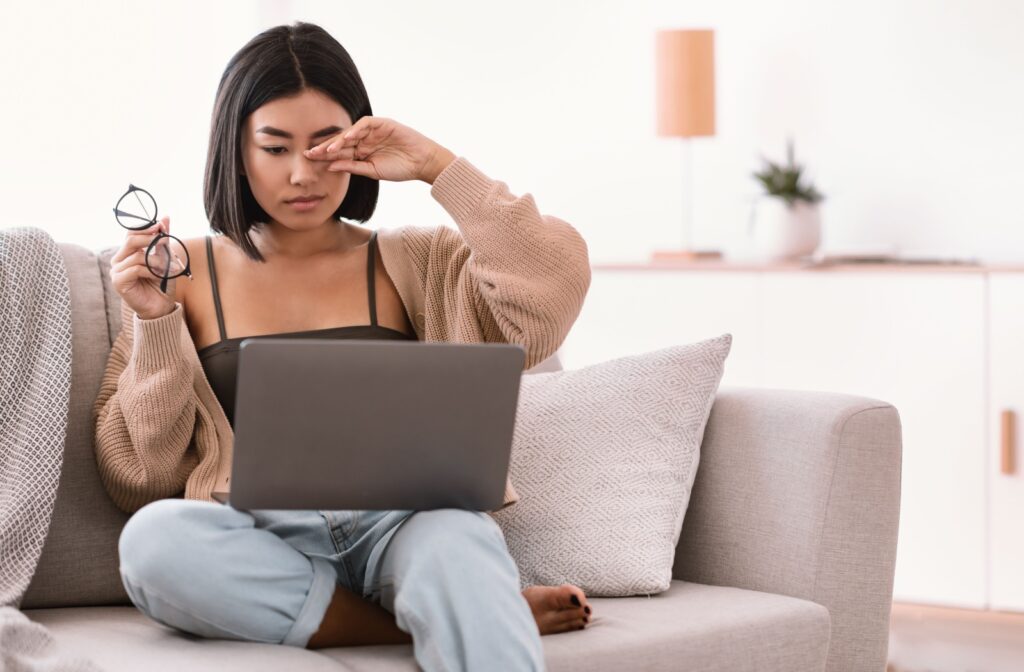 Young woman sitting in front of her computer holding her glasses in one hand and rubbing her eye with her other hand