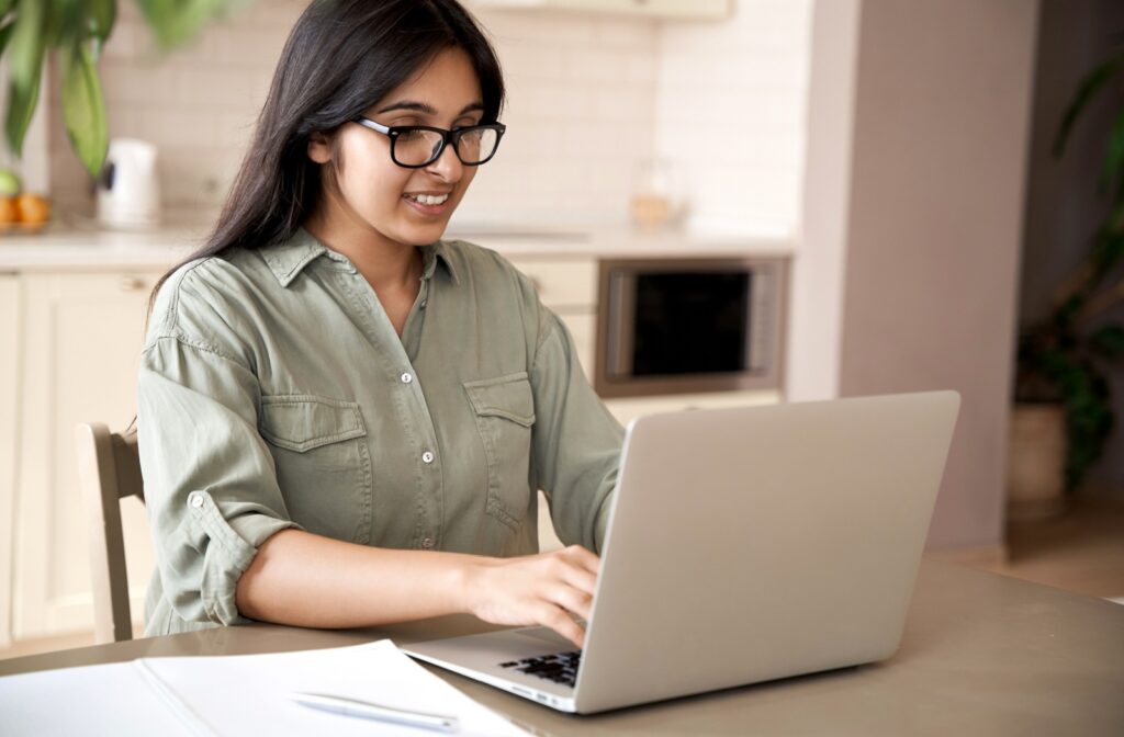Young person wearing glasses and sitting at a table using a laptop with notes next to them.