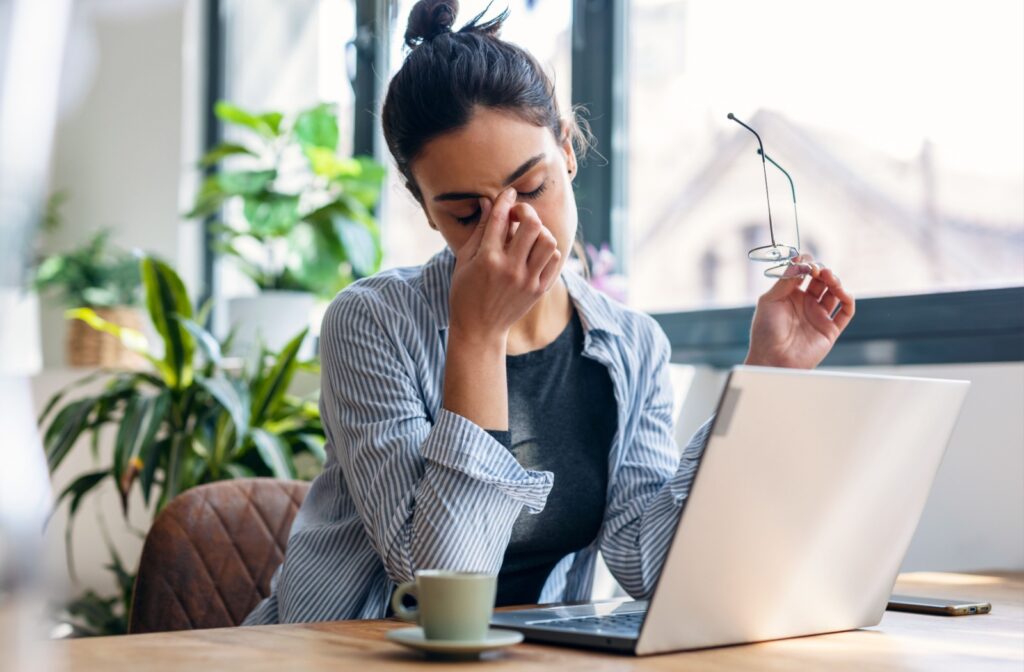 A person sitting in front of a laptop holding their glasses in one hand and rubbing their eyes with their other hand