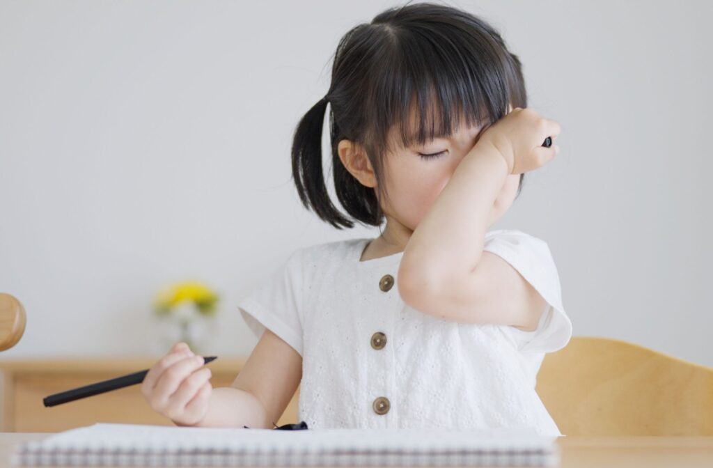 A young child rubs their eye as they colour on a piece of paper